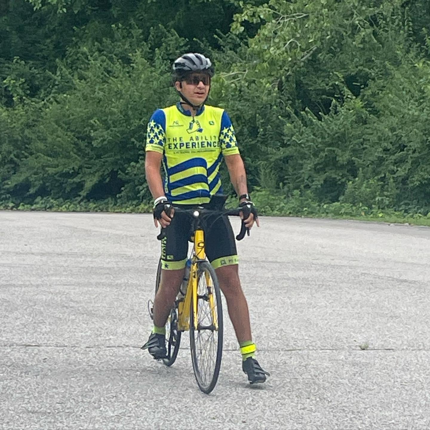 Cyclist wearing helmet, sunglasses, and a blue and yellow jersey that reads The Ability Experience standing with one foot on a yellow bike on a paved road.