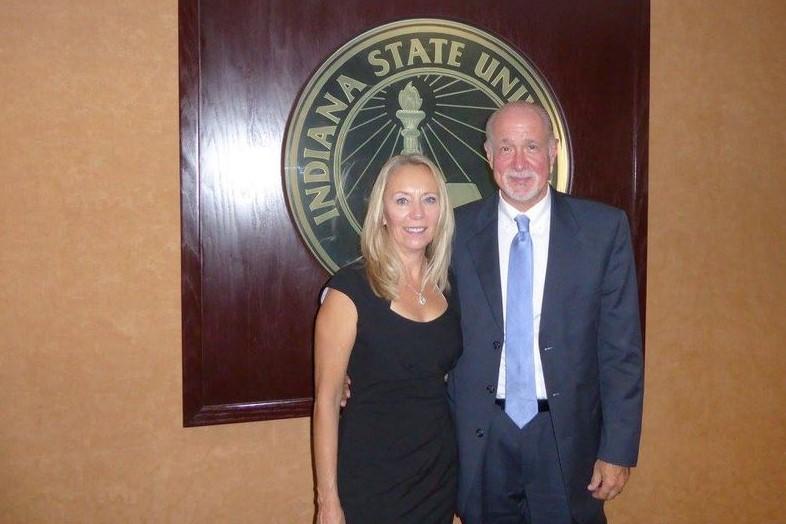 A woman in a black dress and a man in a suit stand together in front of an Indiana State University emblem on a wall.