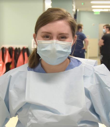 Young woman wearing a surgical mask and hospital gown in a clinical setting with medical staff blurred in the background.