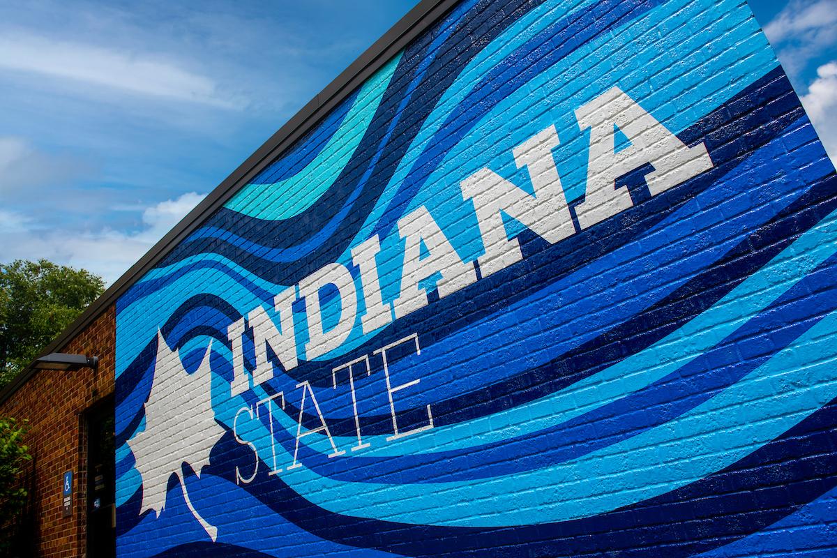 Blue brick wall mural with white text reading Indiana State and a white stylized leaf logo on a building exterior under a partly cloudy sky.