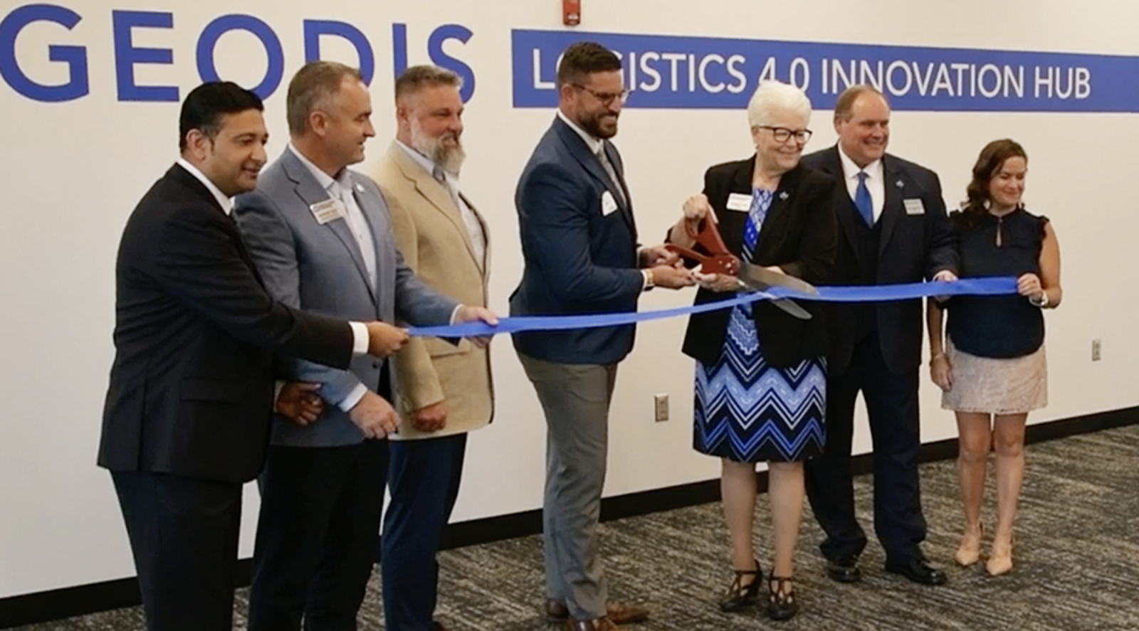 Seven people in business attire cut a blue ribbon at GEODIS Logistics 4.0 Innovation Hub during a formal ribbon-cutting event.