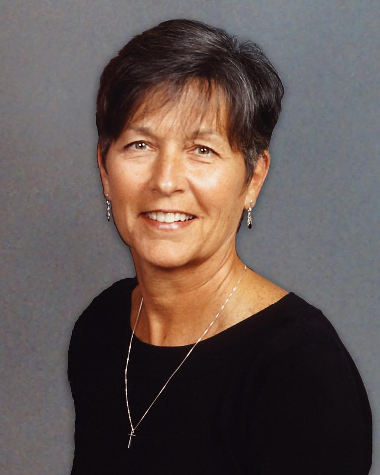 Smiling woman with short dark hair wearing a black top, a silver cross necklace, and dangling earrings against a plain gray background.