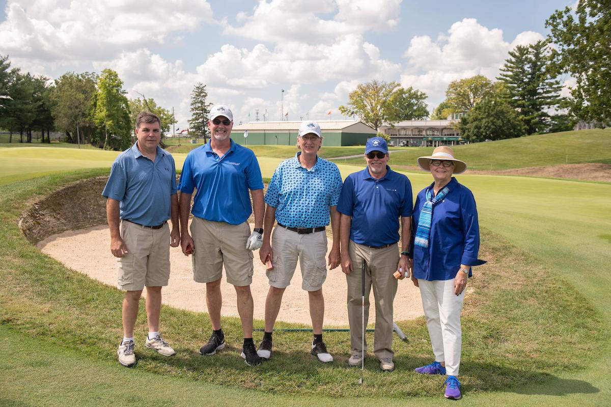 Five adults standing on a golf course near a sand bunker, dressed in casual golf attire, with trees and a building in the background under a partly cloudy sky.