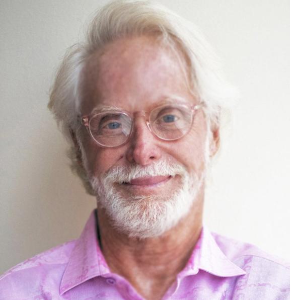 Elderly man with white hair and beard, wearing round glasses and a light pink collared shirt, smiling gently against a plain light background.