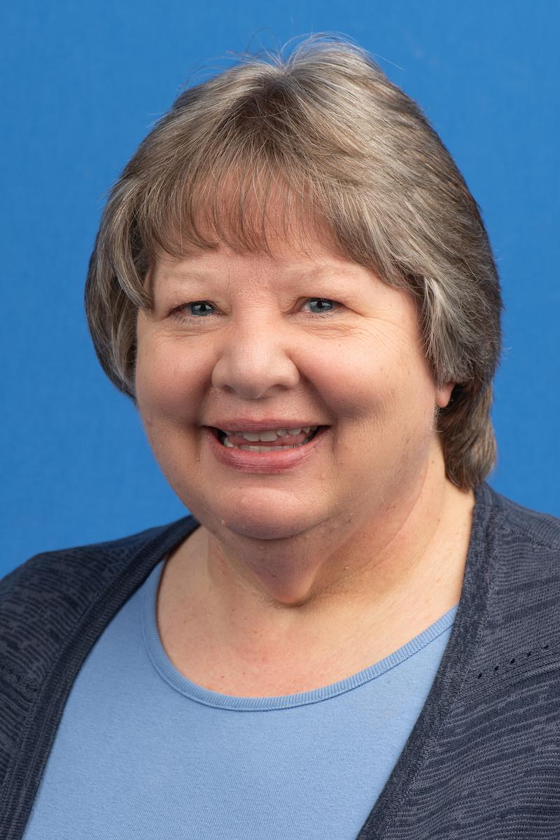Smiling middle-aged woman with short light brown hair wearing a blue top and dark cardigan, against a solid blue background.