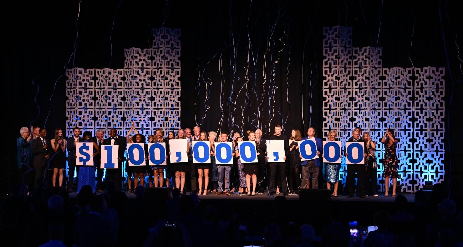 Group of diverse people on stage holding large cards that spell out $100,000,000 with decorative panels and streamers behind them.