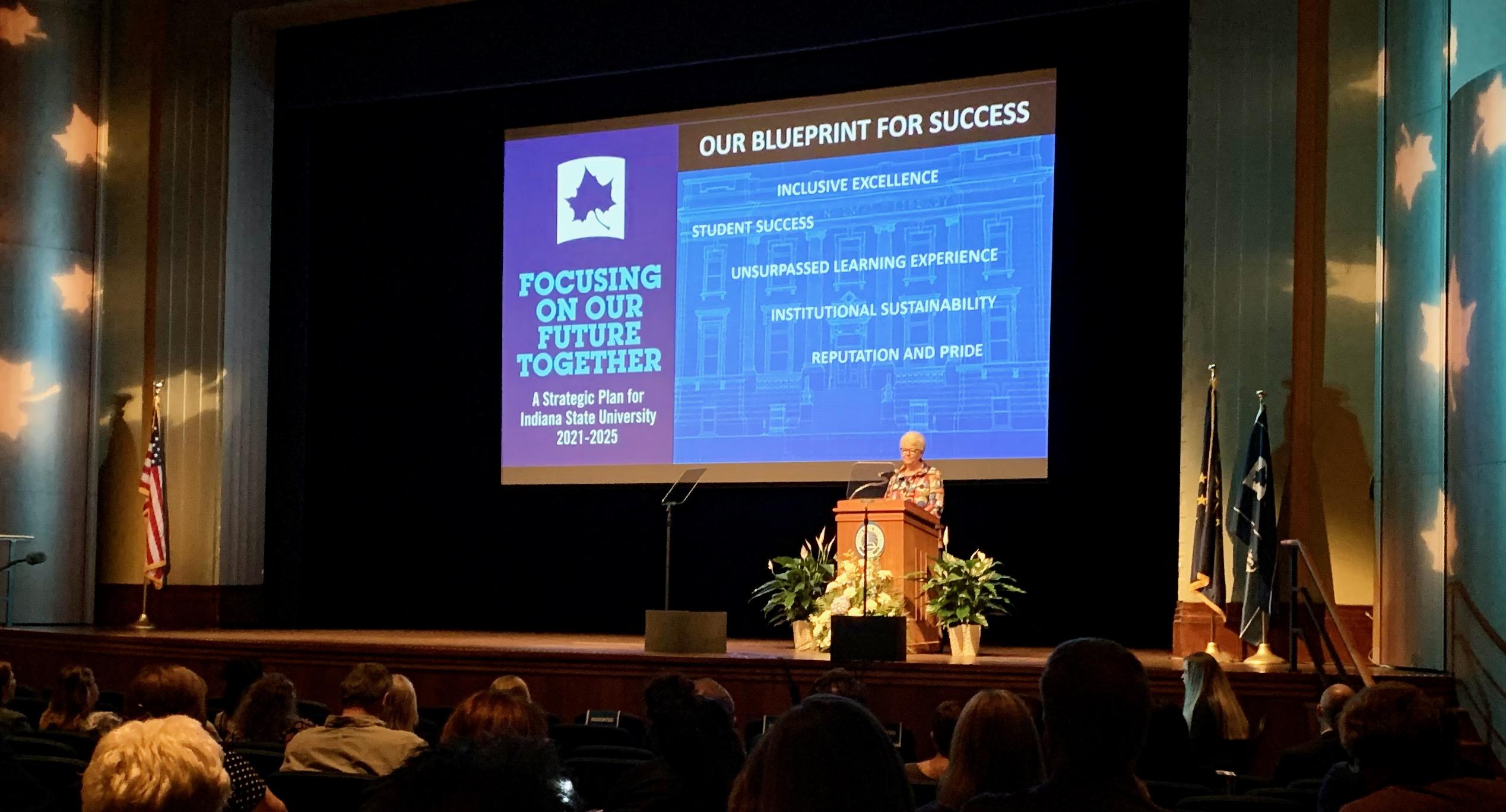 University president standing at podium on stage with large screen behind showing strategic plan titled Our Blueprint for Success and audience seated in foregro