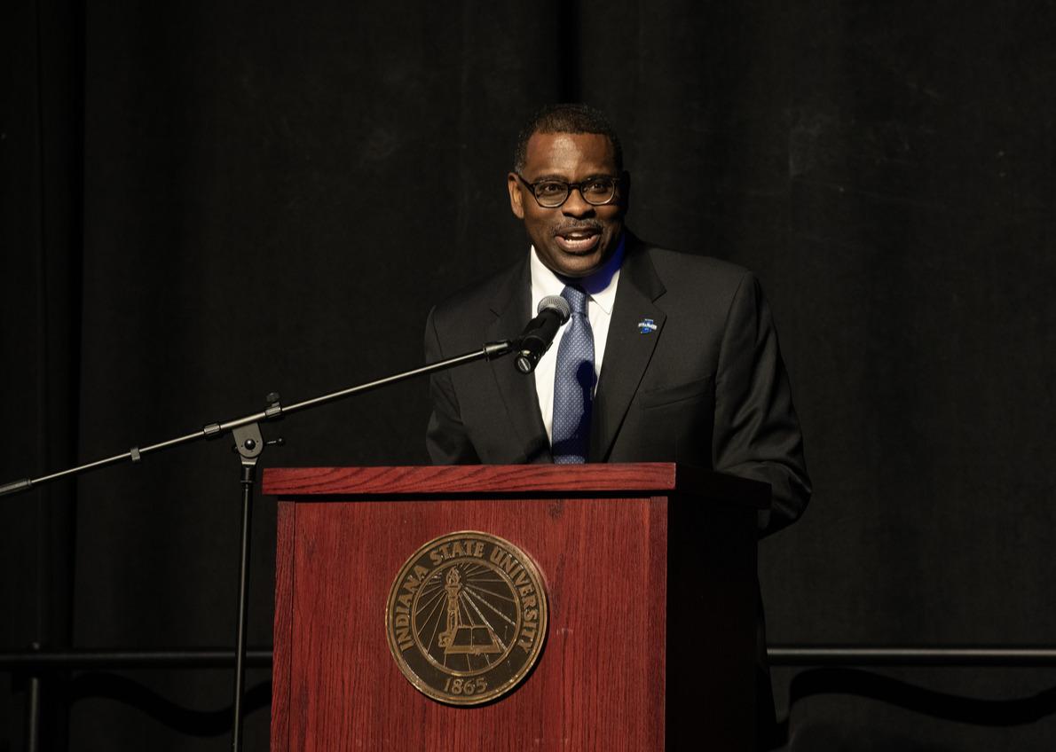 Man in dark suit and blue tie speaking at a wooden podium with Indiana State University emblem, against a black background.