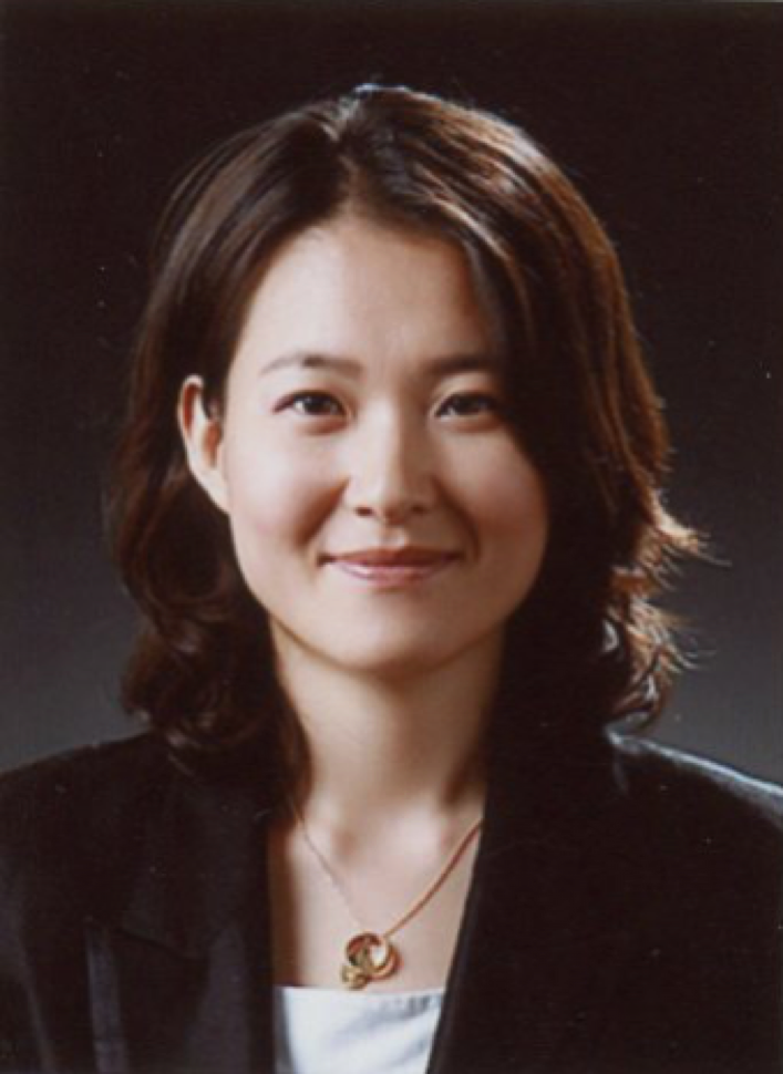 Woman with shoulder-length dark hair wearing a black blazer and white top, smiling against a dark brown background.