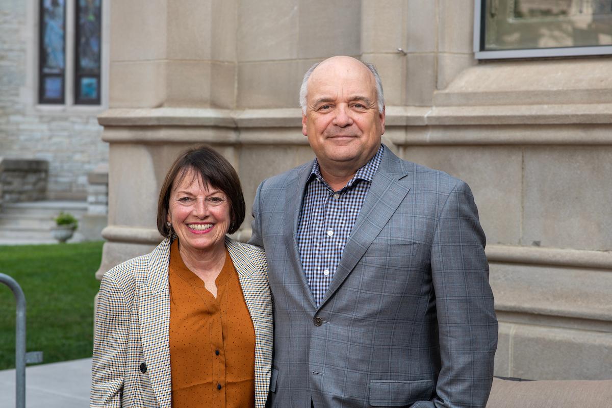 Smiling middle-aged man and woman standing side by side outdoors in front of a stone building, dressed in business casual attire.