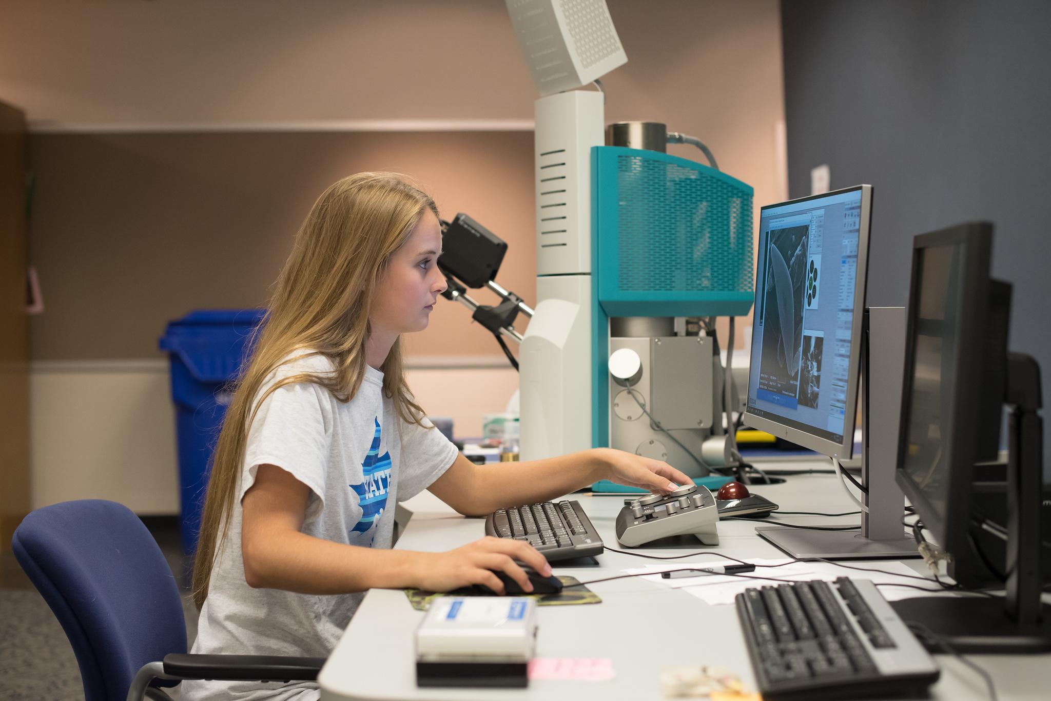 Young woman with long blonde hair using scientific equipment and computer in a lab, analyzing images displayed on a monitor.