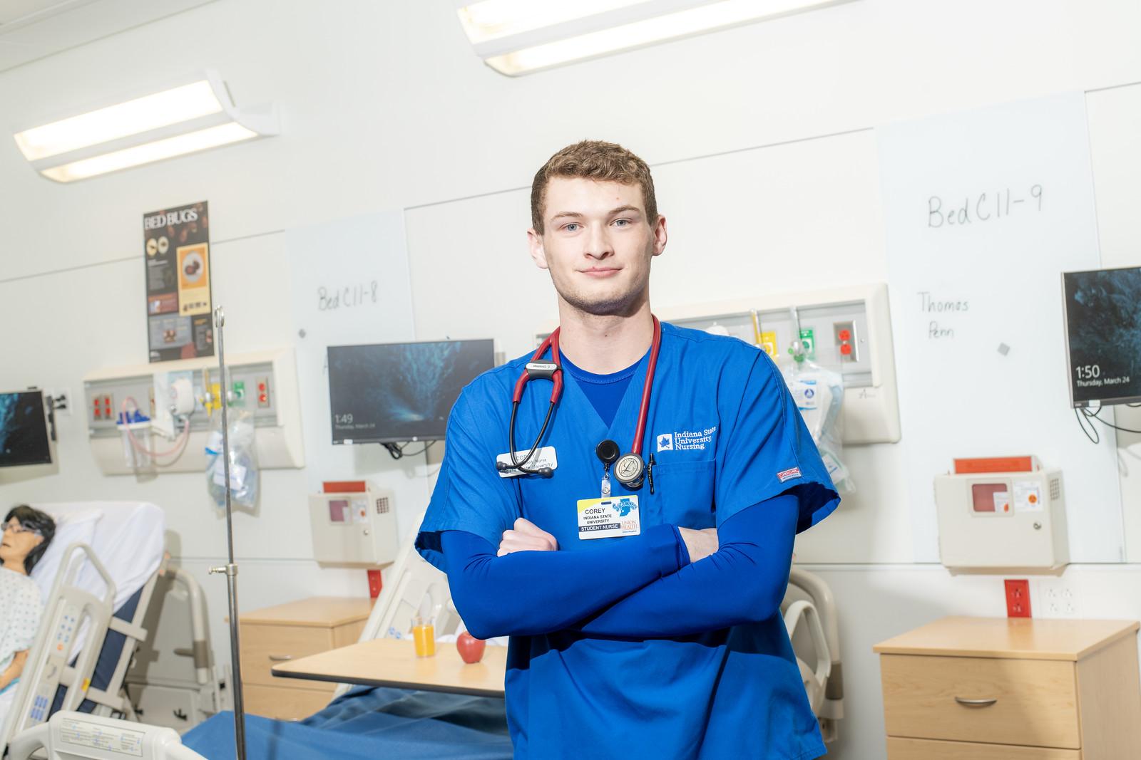 Young male nursing student in blue scrubs with stethoscope stands in hospital room with beds and medical equipment behind him.