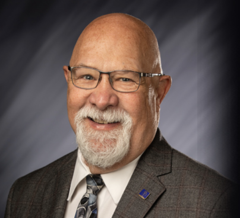Elderly man with glasses, white beard, and mustache wearing a dark plaid suit, white shirt, patterned tie, and a small blue lapel pin against a blurred dark bac