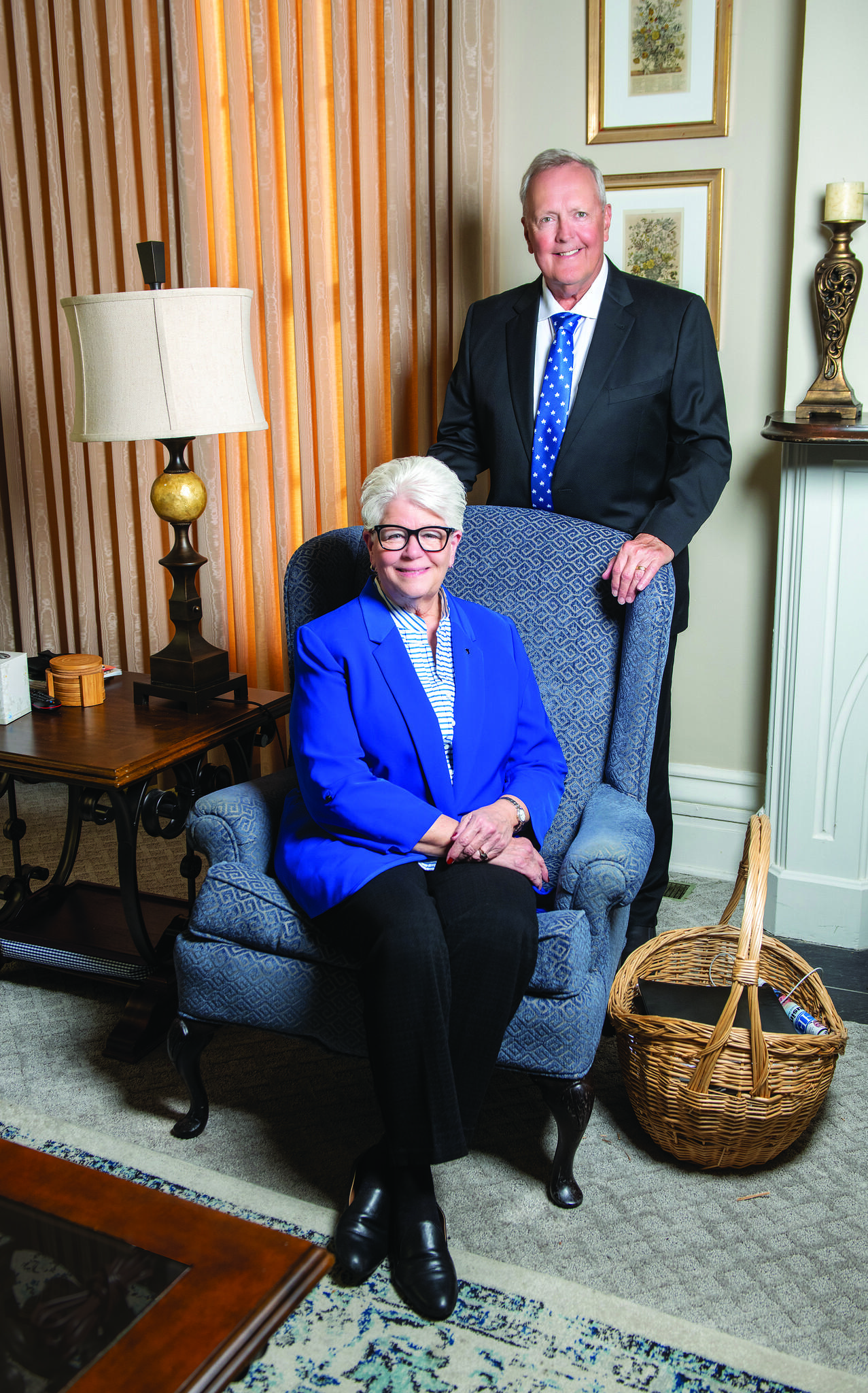 Smiling older couple in formal attire, woman seated in blue armchair wearing blue blazer and glasses, man standing behind her in a suit with blue tie.