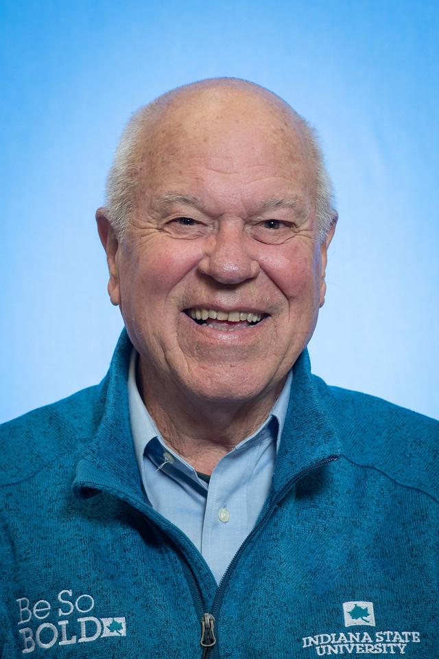 Smiling elderly man with short white hair wearing a blue zip-up jacket with "Be So Bold" and Indiana State University logos on a light blue background.