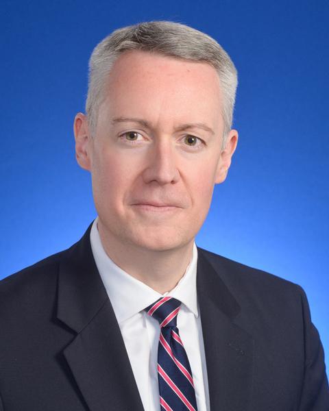Middle-aged man with short gray hair wearing a dark suit, white shirt, and striped tie against a blue background.