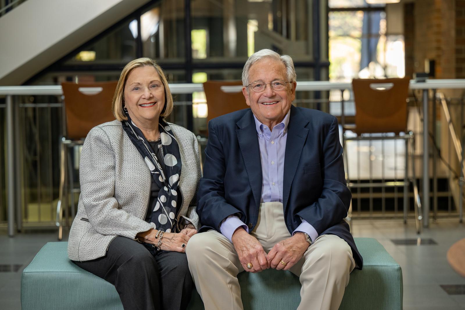 An older man wearing glasses, a navy blazer, and beige pants sits next to a woman in a gray blazer, black pants, and a black and white scarf on a teal bench ind