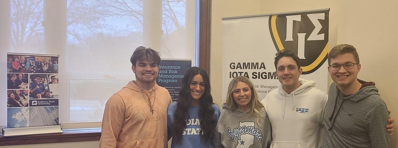 Five Indiana State University students stand smiling indoors between two banners, one for Insurance and Risk Management Program and one for Gamma Iota Sigma.