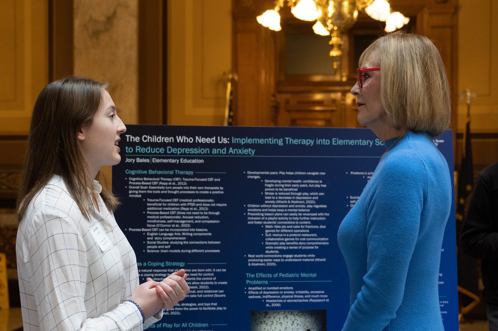 Young woman explains a research poster on therapy for children with depression to an older woman wearing red glasses in a formal indoor setting.