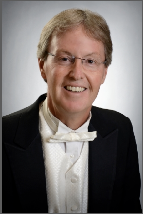 Smiling middle-aged man wearing glasses, a white bow tie, white vest, and black tuxedo jacket against a plain light background.