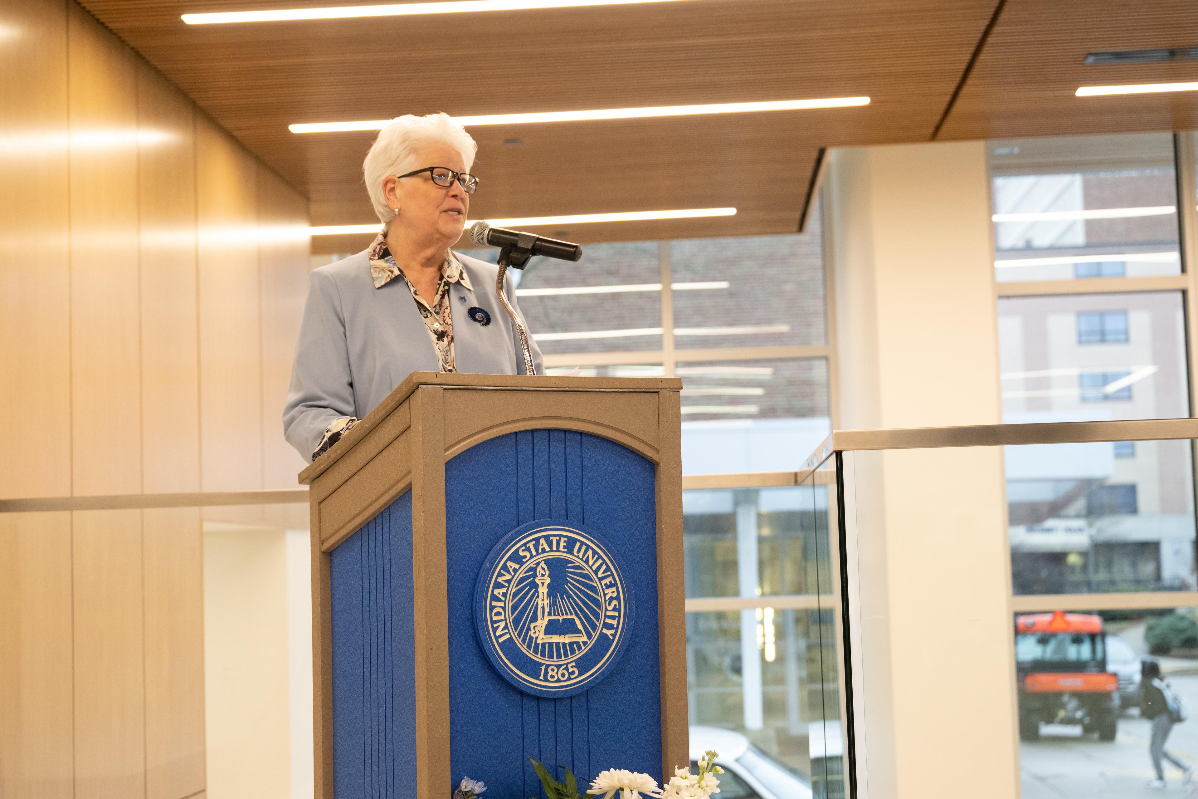 Woman with white hair and glasses speaking at a podium with Indiana State University seal in a modern indoor space with large windows and wood-paneled ceiling.