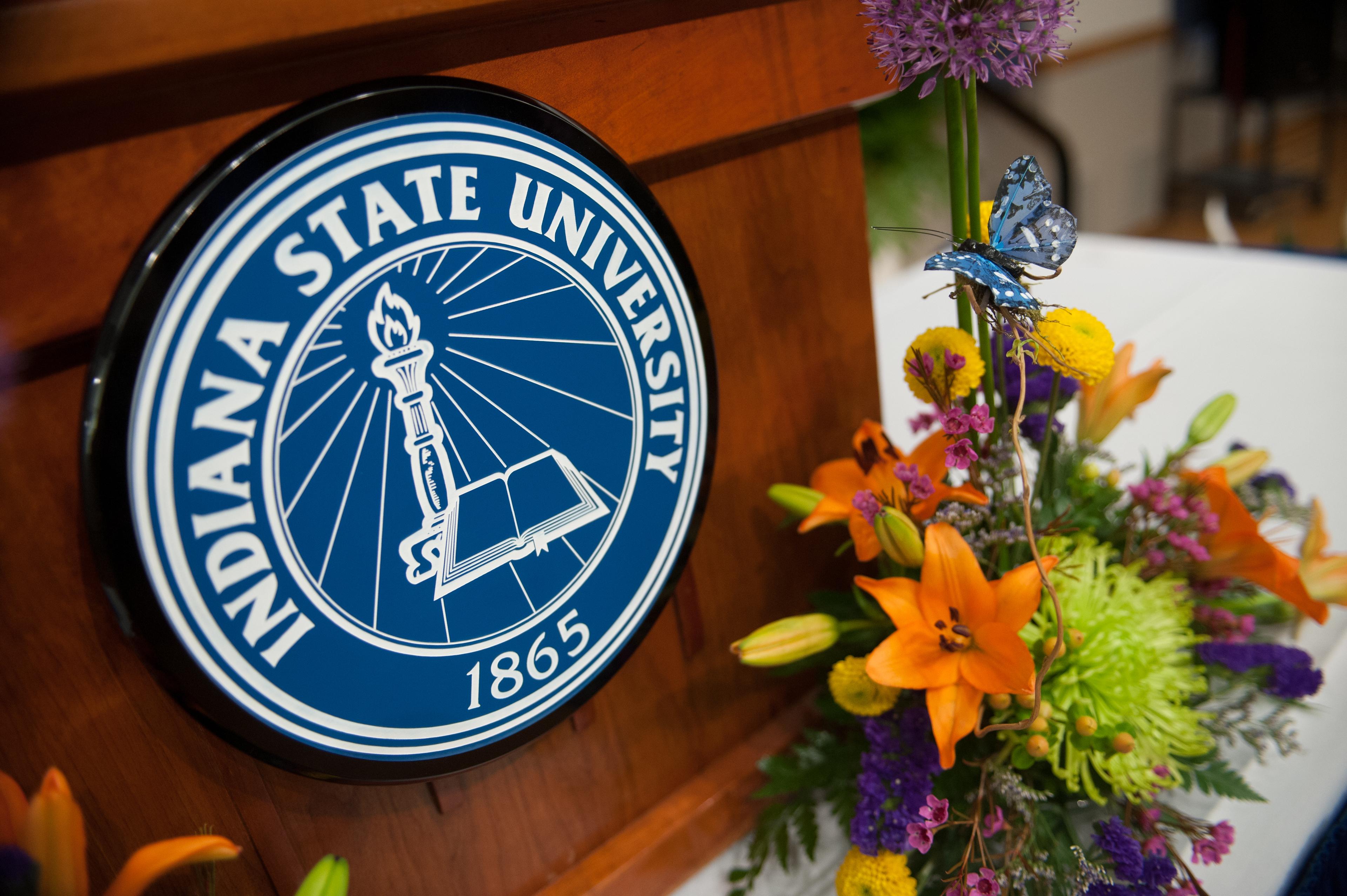 Indiana State University emblem on wooden podium next to a colorful floral arrangement with orange lilies and a decorative blue butterfly.