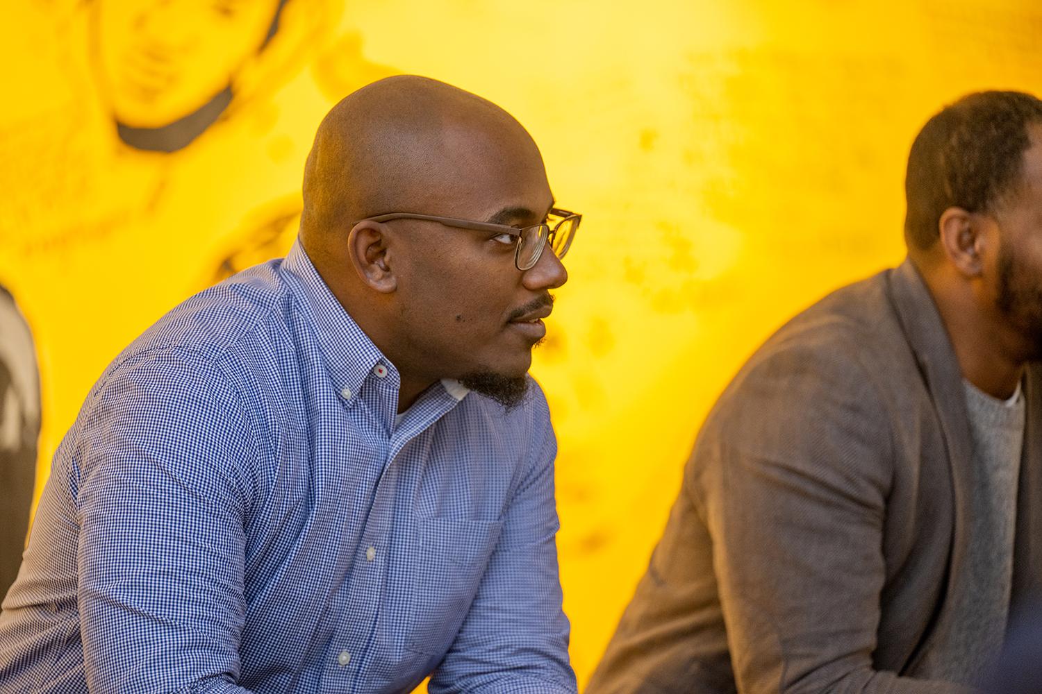 Two men sit in front of a bright yellow wall with a partial black and white portrait visible behind them.