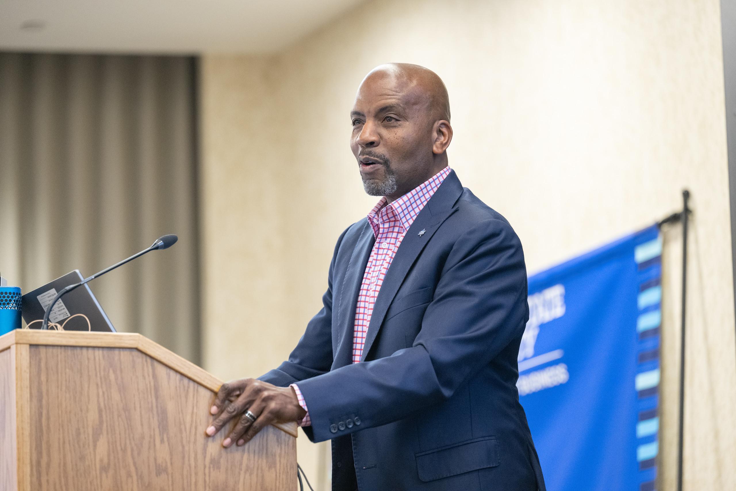 Man in a navy blazer and checkered shirt speaking at a wooden podium with a microphone and laptop, blue event banner in the background.