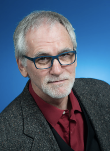 Older man with gray hair, beard, and mustache wearing blue glasses, a red shirt, black vest, and dark blazer against a blue background.