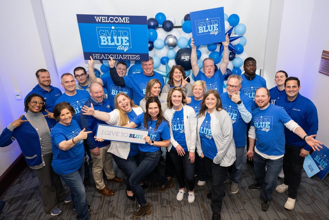 Group of Indiana State University supporters wearing blue Give to Blue Day shirts, holding signs and celebrating in a decorated room.