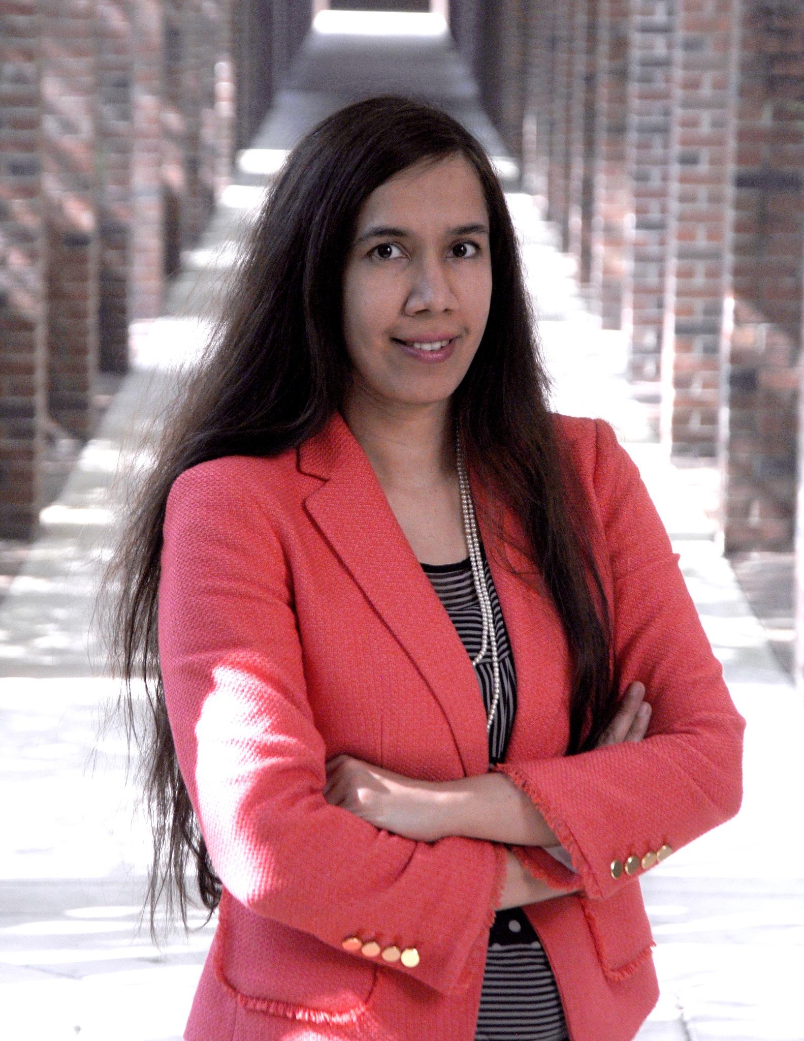 Woman with long dark hair wearing a coral blazer and pearl necklace stands with arms crossed in a sunlit corridor with brick arches.