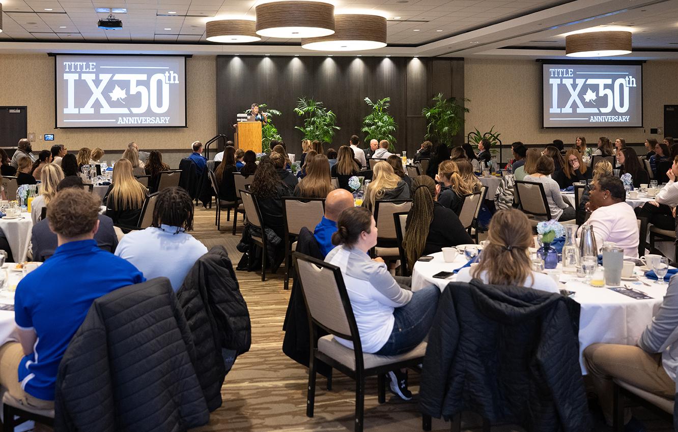 Audience seated at round tables listens to a speaker at a podium in a conference room with two screens displaying Title IX 50th Anniversary.