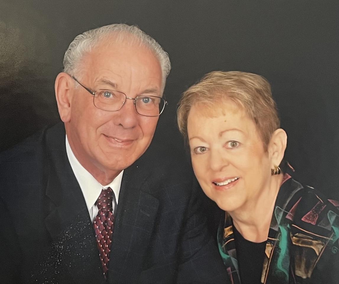 Older man wearing glasses, dark suit, white shirt, red patterned tie next to woman with short hair wearing a colorful patterned jacket, dark background.