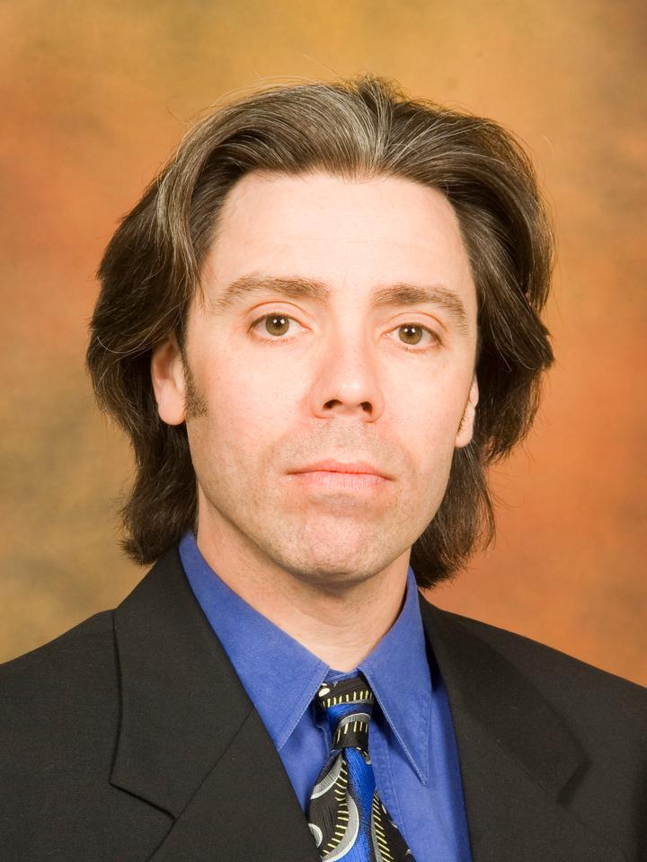 Middle-aged man with medium-length brown hair wearing a black suit, blue shirt, and patterned tie against a warm brown background.