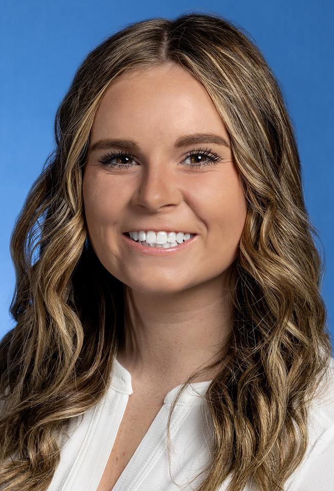 Young woman with wavy light brown hair wearing a white top smiles against a blue background.