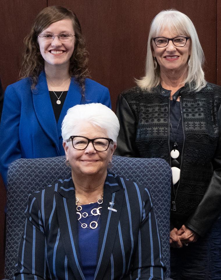 Three women smiling, one seated wearing a blue striped blazer, two standing behind her, one in a blue blazer and the other in a black jacket with white hair.