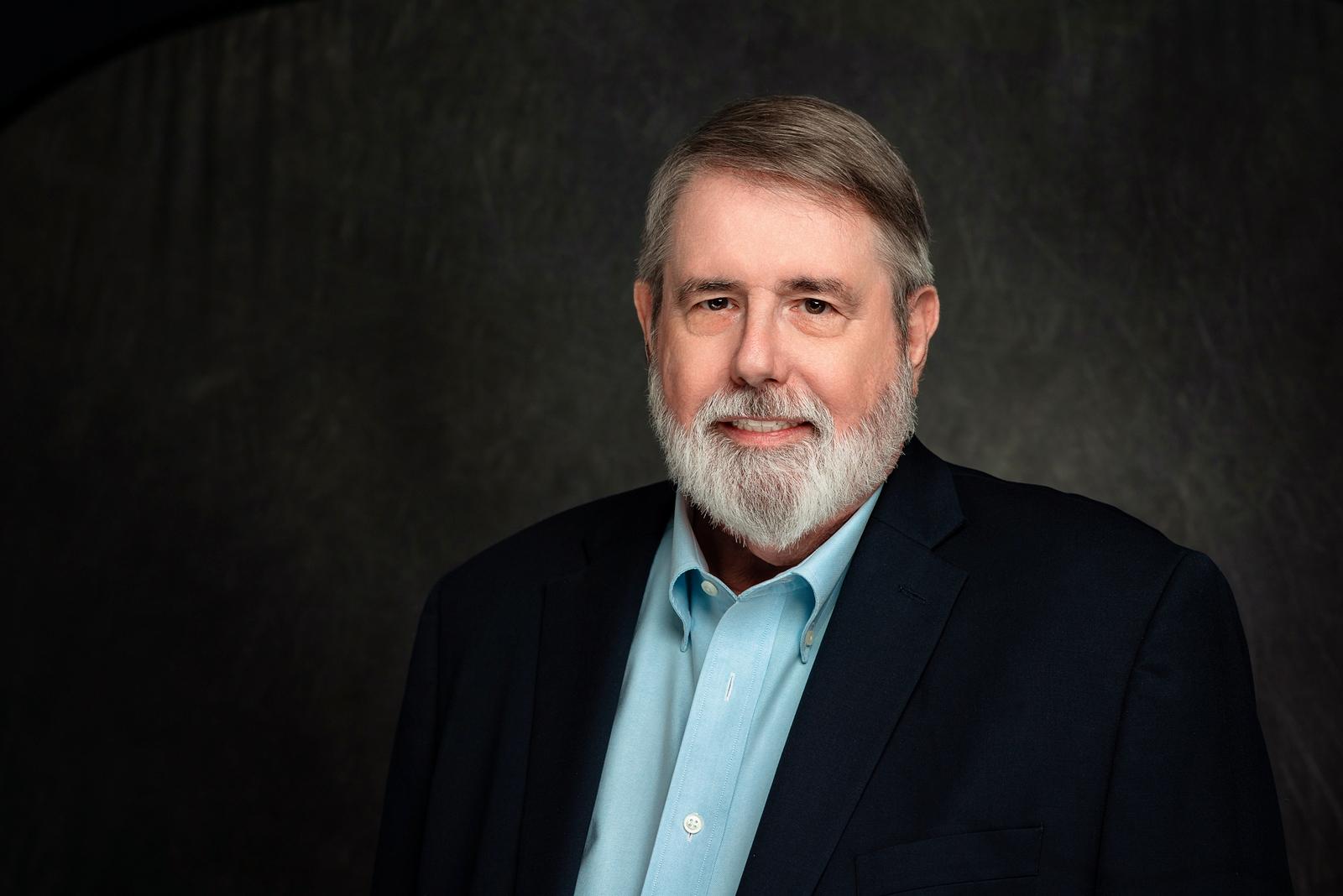 Middle-aged man with gray hair and beard wearing a light blue shirt and dark blazer against a dark gray background.