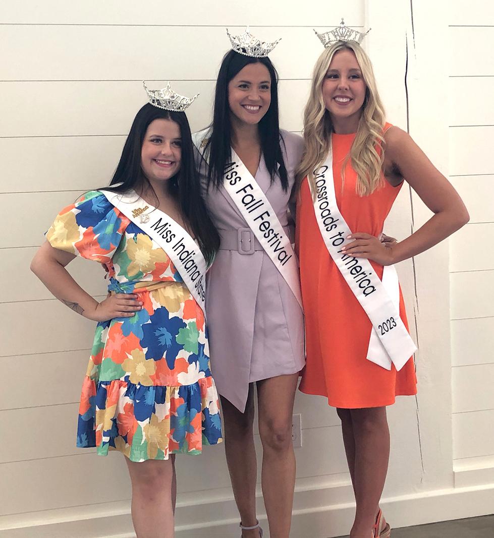 Three smiling women in colorful dresses and crowns wearing Miss Indiana contestant sashes posing in front of a white wall.
