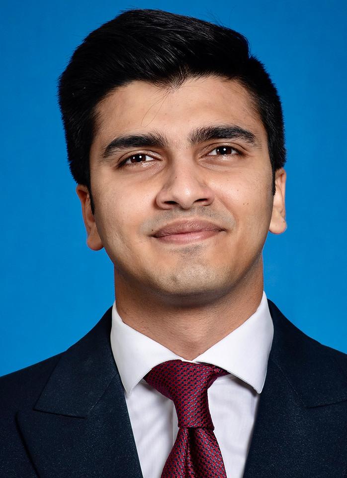Young man with dark hair wearing a dark suit, white shirt, and red patterned tie against a blue background.