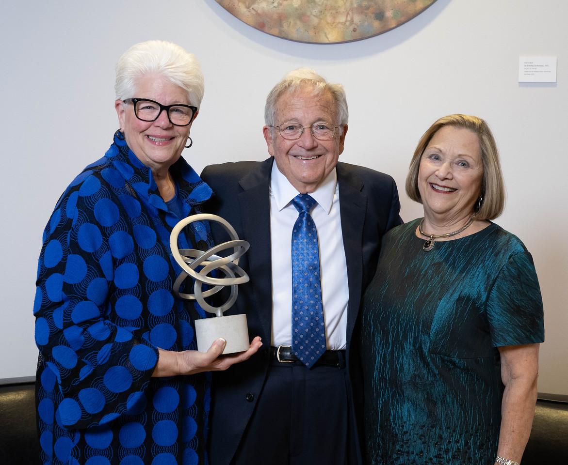 Three people smiling indoors, one woman on the left holding a modern abstract silver sculpture, all dressed in formal attire.