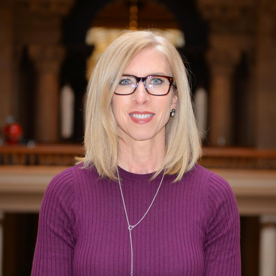 Blonde woman with straight hair wearing glasses, a purple ribbed sweater, and a silver necklace standing indoors with blurred architectural background.