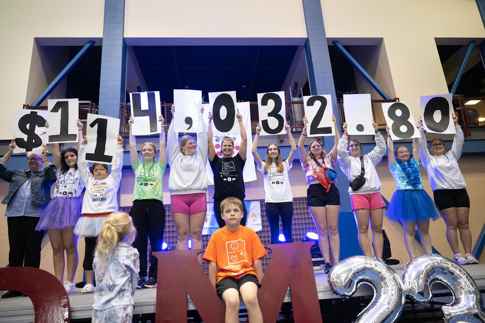 Group of people standing on stage holding large signs showing $114,032.80 with two children seated and standing below them.