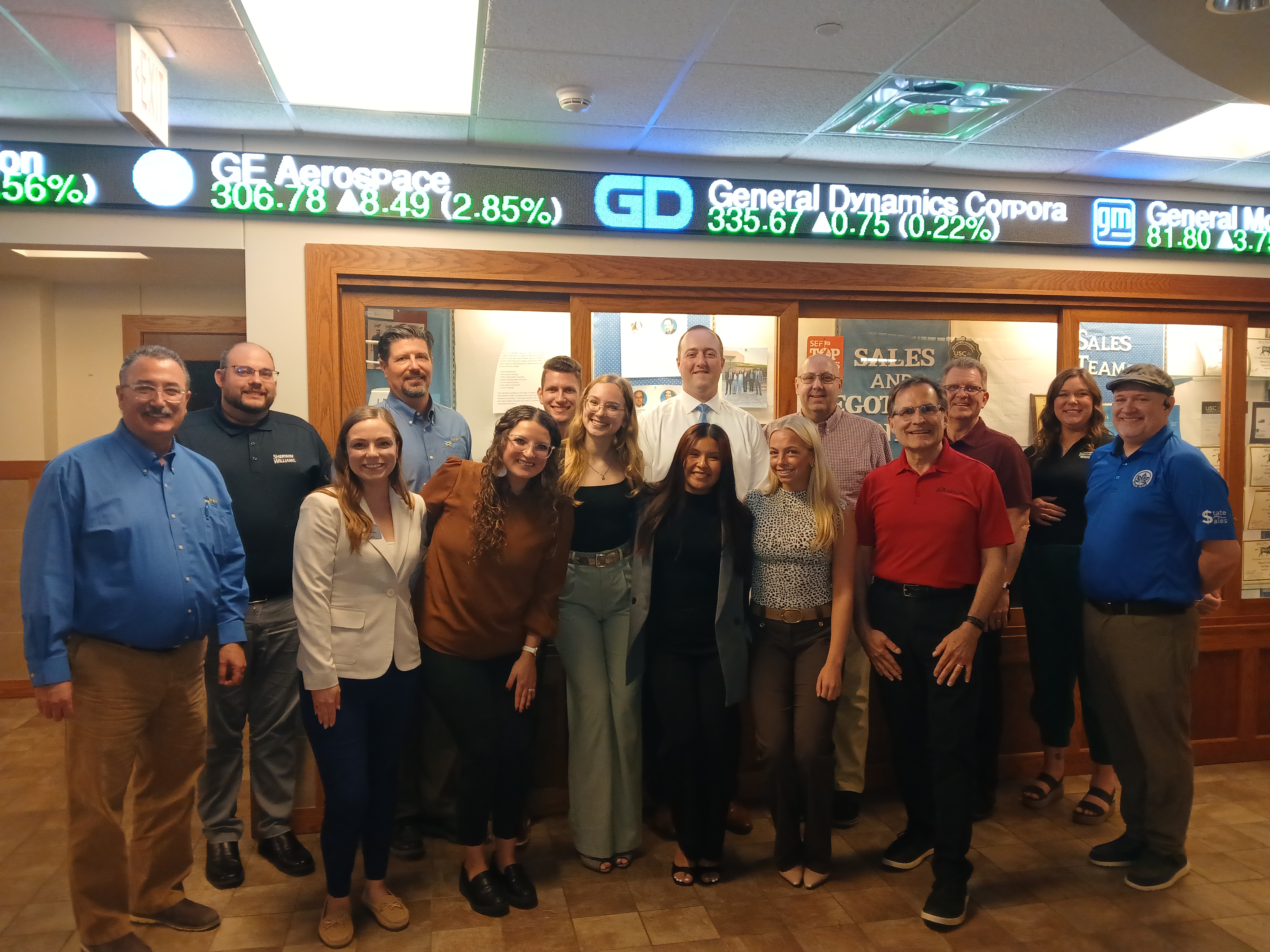 Group of adults and students standing closely together in a hallway with stock ticker and sales team signs visible in the background.