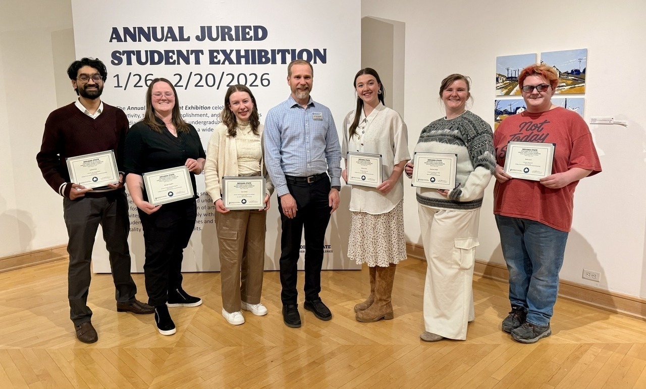 Seven people stand in a row inside a gallery, six holding certificates, with an Annual Juried Student Exhibition sign in the background.