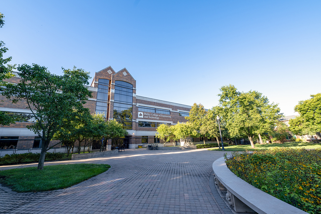 Brick and glass university building labeled Bailey College of Engineering and Technology, surrounded by trees and a curved flowerbed under a clear blue sky.