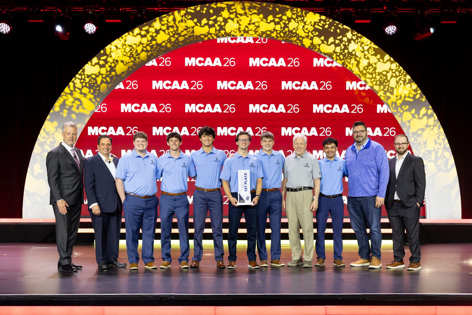 Ten men standing on stage under a large yellow arch in front of a red MCAA 26 backdrop, one holding a 1st place award.