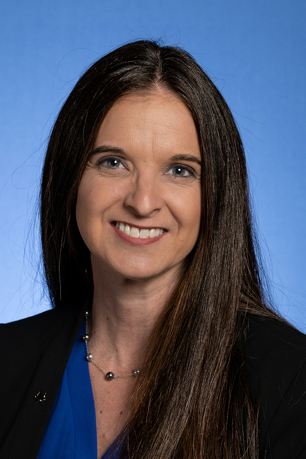Woman with long straight brown hair wearing a blue top, black blazer, and silver necklace smiling against a blue background.