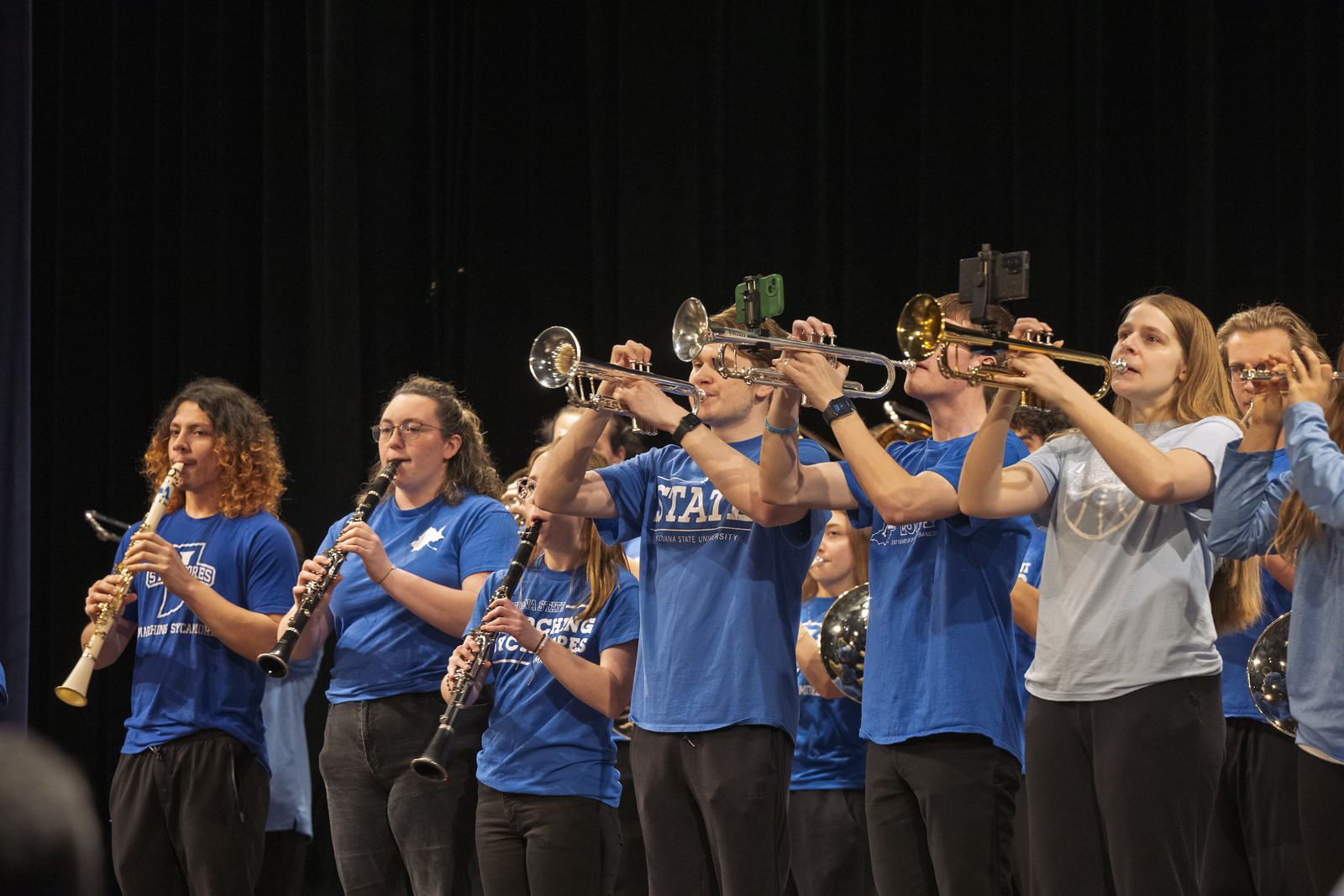 Group of Indiana State University band members in blue shirts playing clarinets and trumpets on stage against a black curtain background.