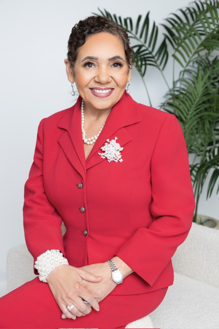 Woman with short curly hair wearing a red blazer adorned with pearl accessories, sitting and smiling against a white background with green plants.