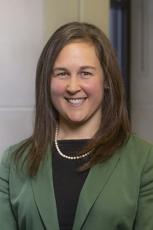 Smiling woman with shoulder-length brown hair, wearing a green blazer, black top, and pearl necklace, standing indoors in front of a wall.